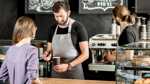 A barista in a gray apron preparing a coffee order for a customer at a modern coffee shop. The customer stands at the counter, while another barista works in the background near an espresso machine. The shop has a cozy, artisanal atmosphere with chalkboard menus in the background.