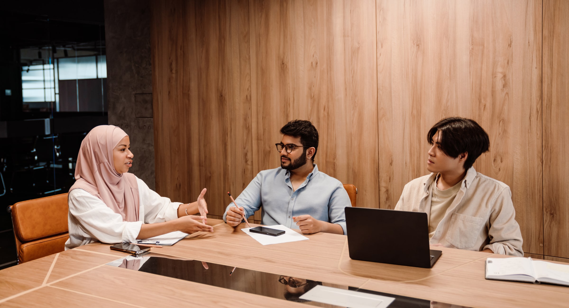 Three colleagues engage in a discussion during a team meeting in a modern office with wood-paneled walls. The group consists of a woman wearing a light pink hijab, who is speaking and gesturing with her hands, a man with glasses holding a pencil, and a third person with medium-length hair using a laptop. The table is equipped with notebooks, a smartphone, and a laptop, indicating a professional and collaborative environment.
