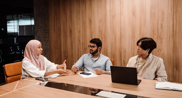 Three colleagues engage in a discussion during a team meeting in a modern office with wood-paneled walls. The group consists of a woman wearing a light pink hijab, who is speaking and gesturing with her hands, a man with glasses holding a pencil, and a third person with medium-length hair using a laptop. The table is equipped with notebooks, a smartphone, and a laptop, indicating a professional and collaborative environment.