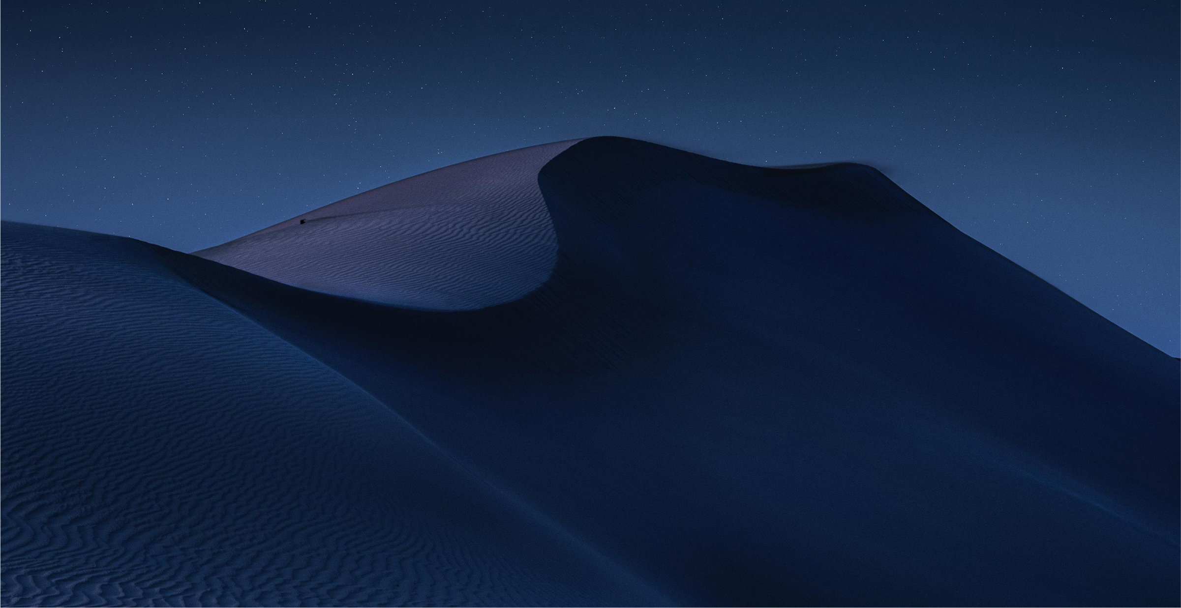 A serene nighttime landscape of desert dunes, softly illuminated under a clear, starry sky. The smooth curves and shadows of the sand create a dramatic, peaceful scene. The image captures the vastness and beauty of the desert under the calmness of night, evoking a sense of solitude and tranquility.