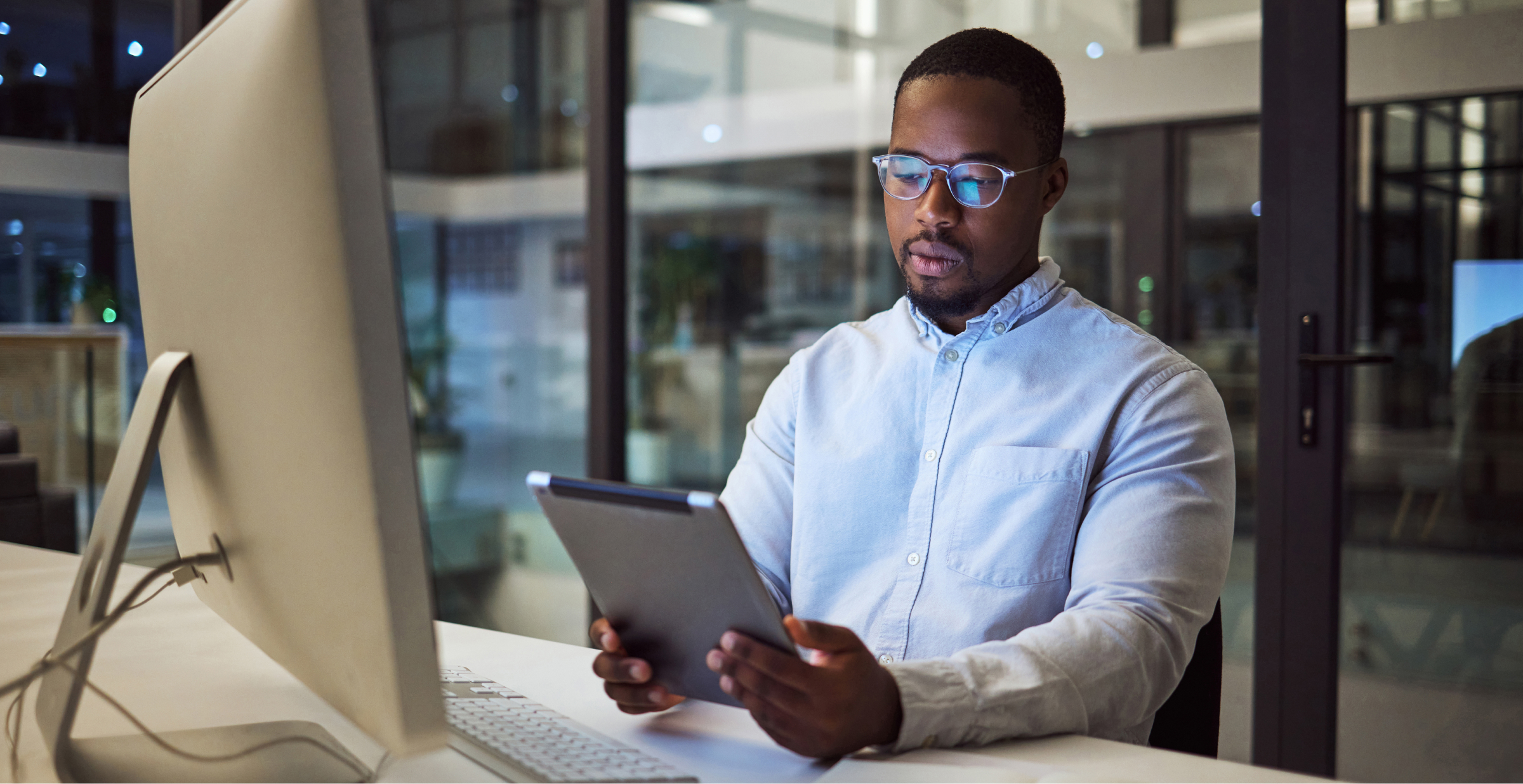 A focused man working late in an office, using a tablet and computer.
