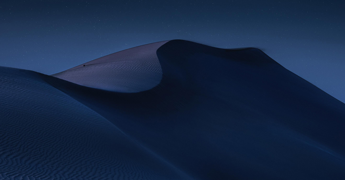 A serene nighttime landscape of desert dunes, softly illuminated under a clear, starry sky. The smooth curves and shadows of the sand create a dramatic, peaceful scene. The image captures the vastness and beauty of the desert under the calmness of night, evoking a sense of solitude and tranquility.