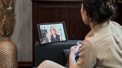 A woman is sitting on a couch, holding her glasses while participating in an online therapy session via a tablet. The tablet screen shows a therapist with pink hair and glasses, speaking and gesturing with her hands. The setting is cozy and intimate, with a vase of dried plants and warm lighting in the background. The scene conveys the convenience and accessibility of mental health support through digital platforms.