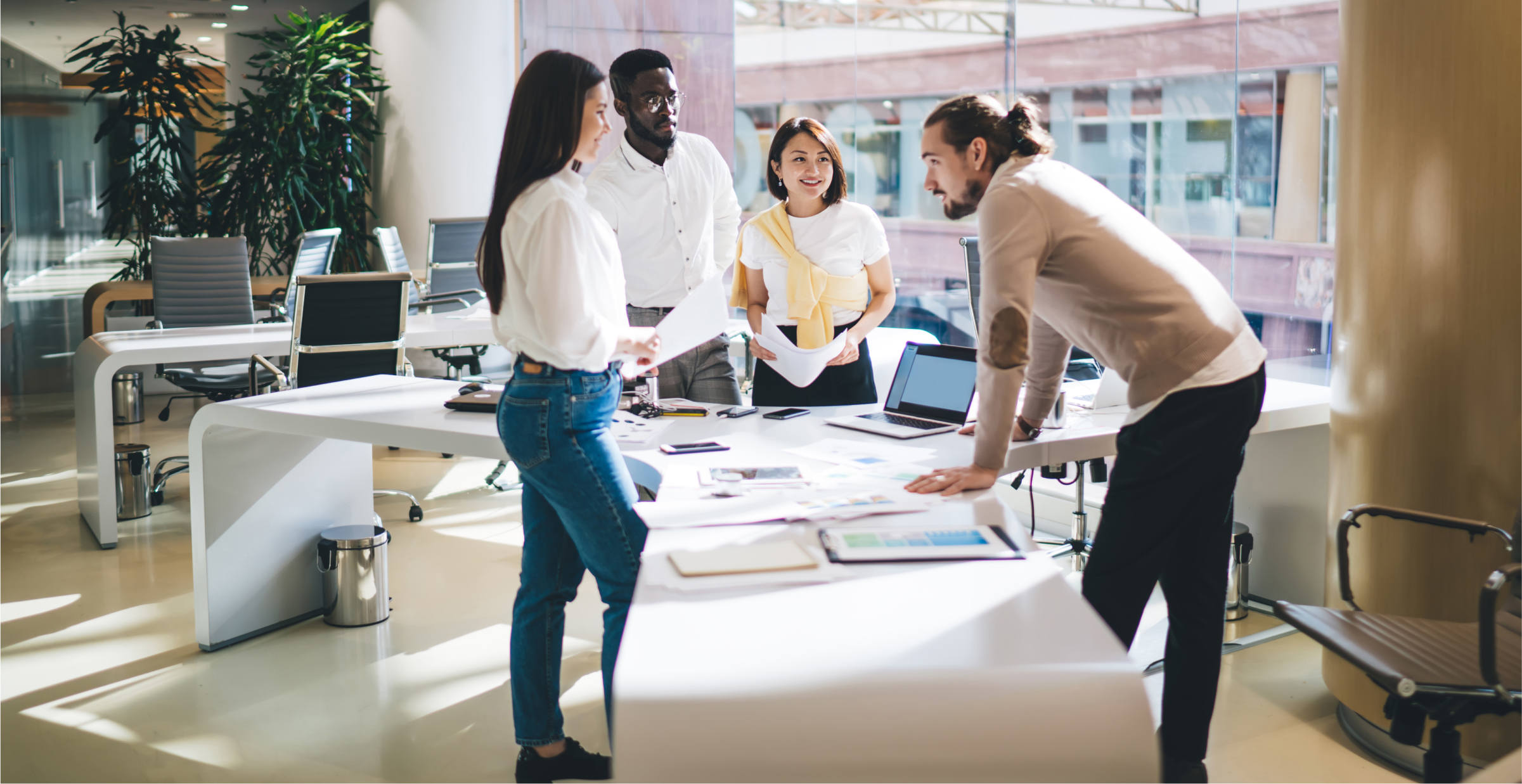 A diverse group of colleagues is gathered around a large table in a modern, bright office. They are engaged in a collaborative discussion, with documents, laptops, and charts spread out on the table. The team appears to be in a brainstorming session, with one member leaning forward and others listening attentively. The office space is spacious, with large windows and contemporary decor, reflecting a professional and dynamic work environment.