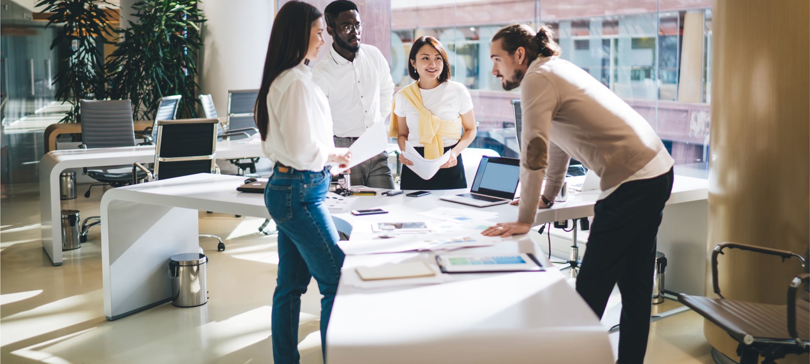 A diverse group of colleagues is gathered around a large table in a modern, bright office. They are engaged in a collaborative discussion, with documents, laptops, and charts spread out on the table. The team appears to be in a brainstorming session, with one member leaning forward and others listening attentively. The office space is spacious, with large windows and contemporary decor, reflecting a professional and dynamic work environment.