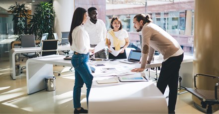 A diverse group of colleagues is gathered around a large table in a modern, bright office. They are engaged in a collaborative discussion, with documents, laptops, and charts spread out on the table. The team appears to be in a brainstorming session, with one member leaning forward and others listening attentively. The office space is spacious, with large windows and contemporary decor, reflecting a professional and dynamic work environment.