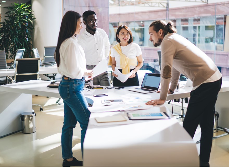 A diverse group of colleagues is gathered around a large table in a modern, bright office. They are engaged in a collaborative discussion, with documents, laptops, and charts spread out on the table. The team appears to be in a brainstorming session, with one member leaning forward and others listening attentively. The office space is spacious, with large windows and contemporary decor, reflecting a professional and dynamic work environment.