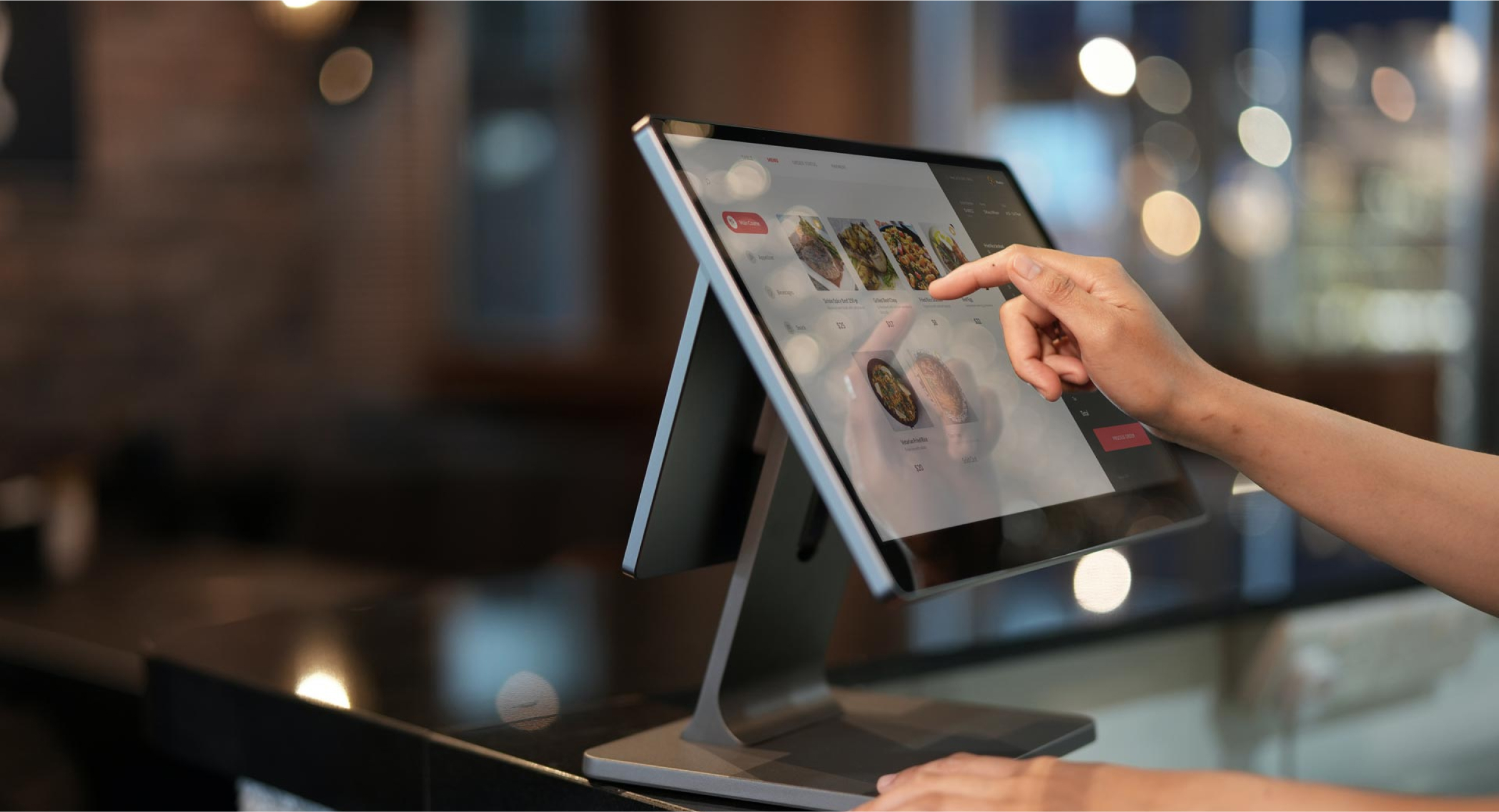 A close-up of a person's hand interacting with a touchscreen tablet at a restaurant or cafe. The screen displays a digital menu with various food items, and the person is selecting an option. The background is softly blurred, creating a focus on the tablet and hand.