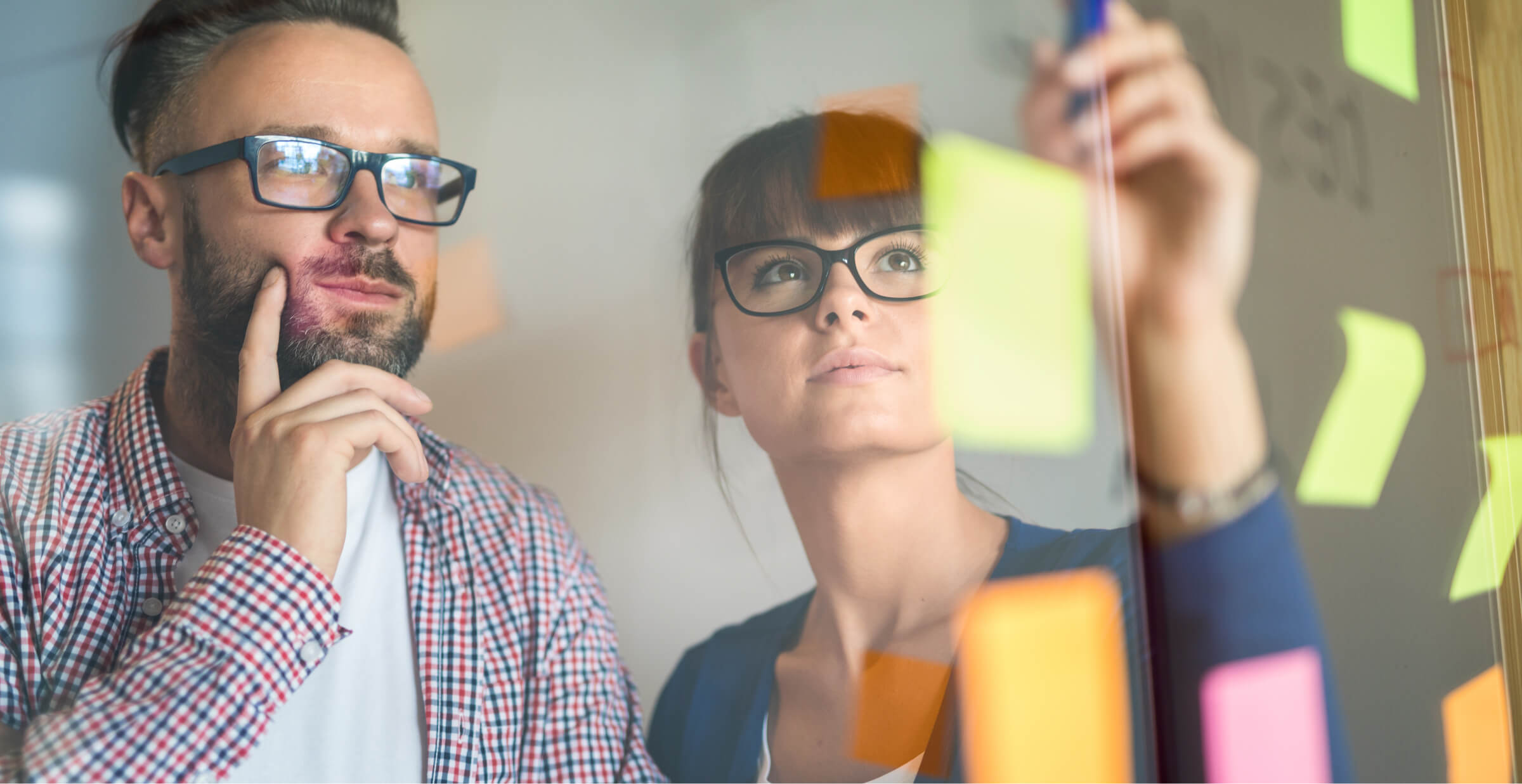 A man and woman, both wearing glasses, are brainstorming in front of a glass wall filled with colorful sticky notes. The man is deep in thought, while the woman is placing or adjusting a note. The scene captures the collaborative and creative process of problem-solving in a modern office environment. The vibrant notes and reflective glass suggest active planning and idea generation.