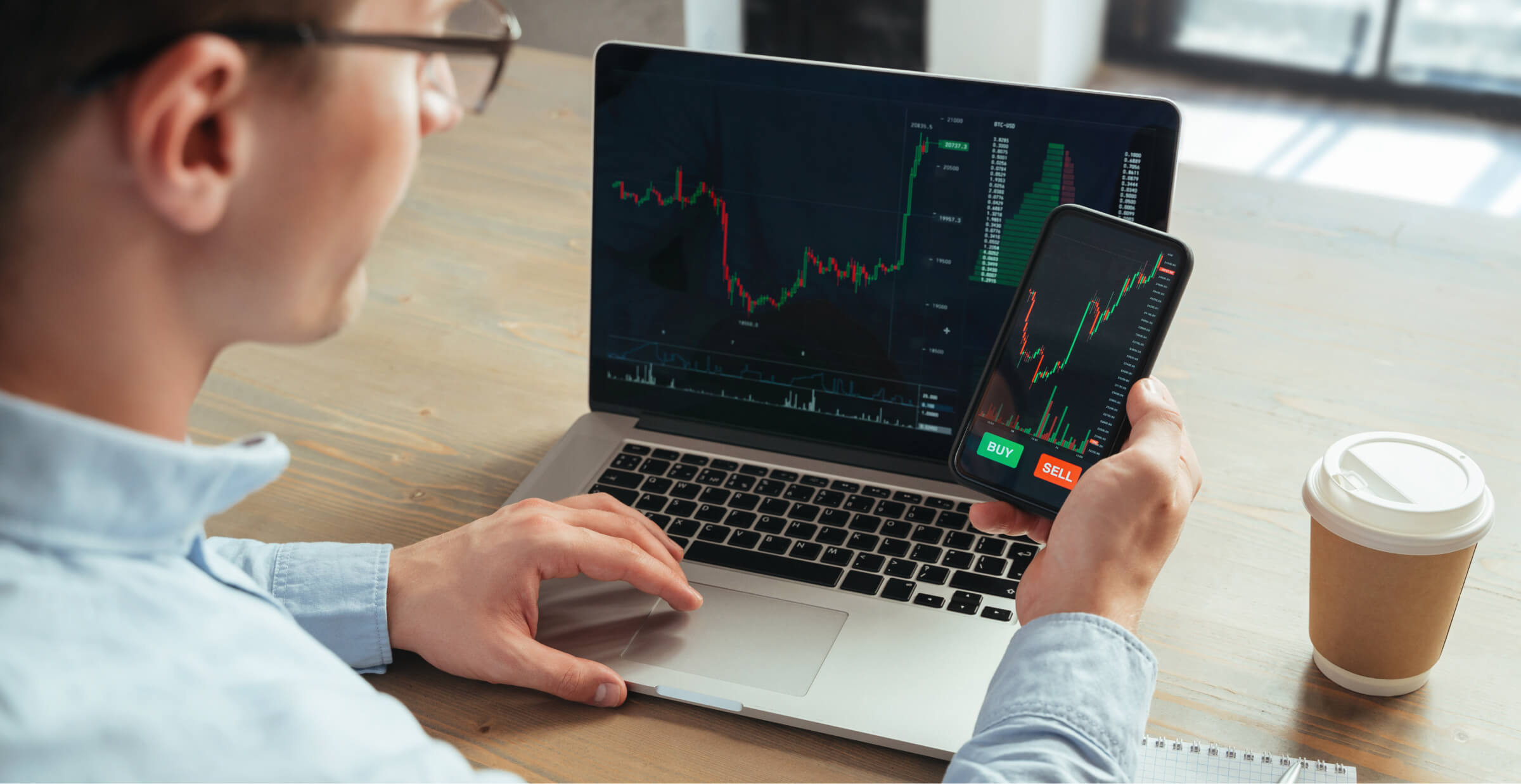 A man is sitting at a desk using both a laptop and a smartphone to monitor stock trading charts. The laptop and phone screens display live financial data with price charts and buy/sell options. A takeaway coffee cup is placed on the desk next to him, indicating a focused and dynamic work environment. The image conveys modern investment management and the use of technology in financial trading.