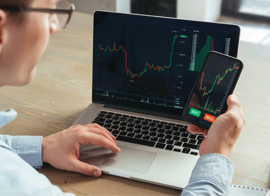 A man is sitting at a desk using both a laptop and a smartphone to monitor stock trading charts. The laptop and phone screens display live financial data with price charts and buy/sell options. A takeaway coffee cup is placed on the desk next to him, indicating a focused and dynamic work environment. The image conveys modern investment management and the use of technology in financial trading.