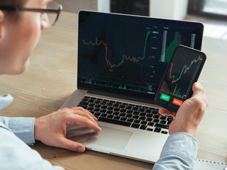 A man is sitting at a desk using both a laptop and a smartphone to monitor stock trading charts. The laptop and phone screens display live financial data with price charts and buy/sell options. A takeaway coffee cup is placed on the desk next to him, indicating a focused and dynamic work environment. The image conveys modern investment management and the use of technology in financial trading.
