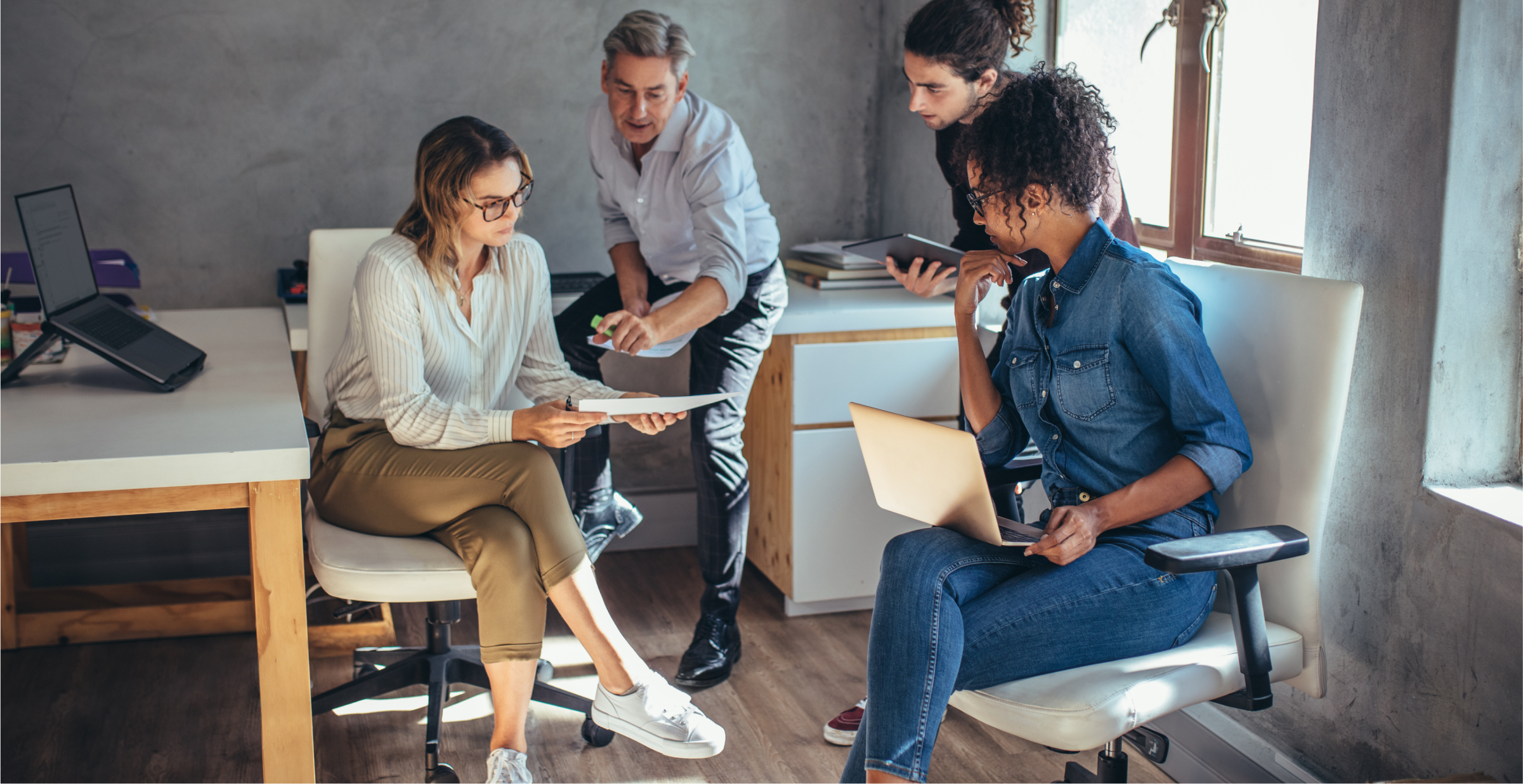 A group of four colleagues is engaged in a collaborative discussion in a modern office setting. One woman holds a document while another reviews it, with two other team members contributing to the conversation. Laptops and tablets are present, indicating a productive work session. The office environment is casual and bright, with natural light streaming through the windows, reflecting a focused and dynamic team effort.