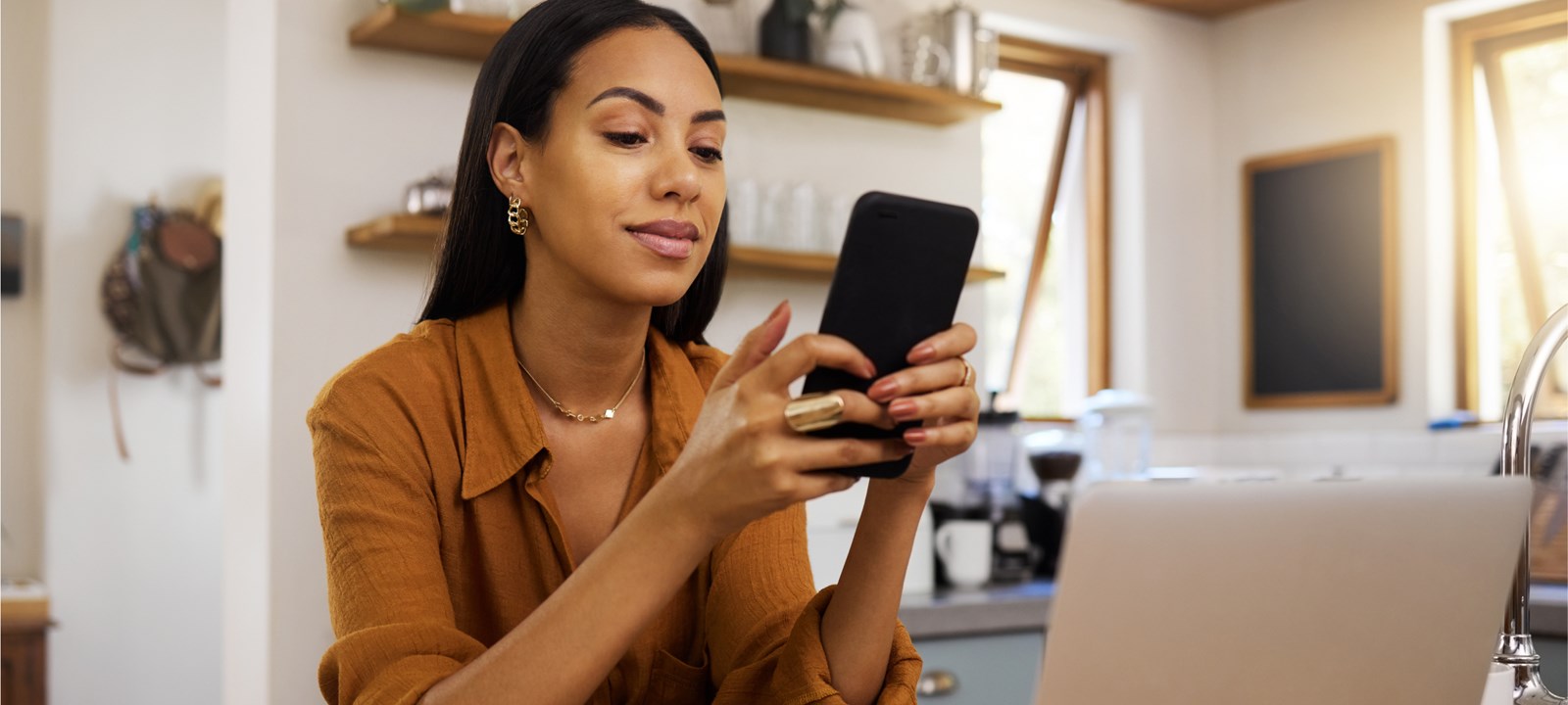 A woman is sitting in a cozy home office, smiling as she uses her smartphone. She appears relaxed and focused, with a laptop in front of her and a modern kitchen in the background. The setting suggests a comfortable and productive work-from-home environment, with natural light streaming through the windows. The image highlights the balance of professional and personal life in a home office setting.
