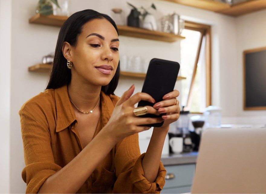 A woman is sitting in a cozy home office, smiling as she uses her smartphone. She appears relaxed and focused, with a laptop in front of her and a modern kitchen in the background. The setting suggests a comfortable and productive work-from-home environment, with natural light streaming through the windows. The image highlights the balance of professional and personal life in a home office setting.
