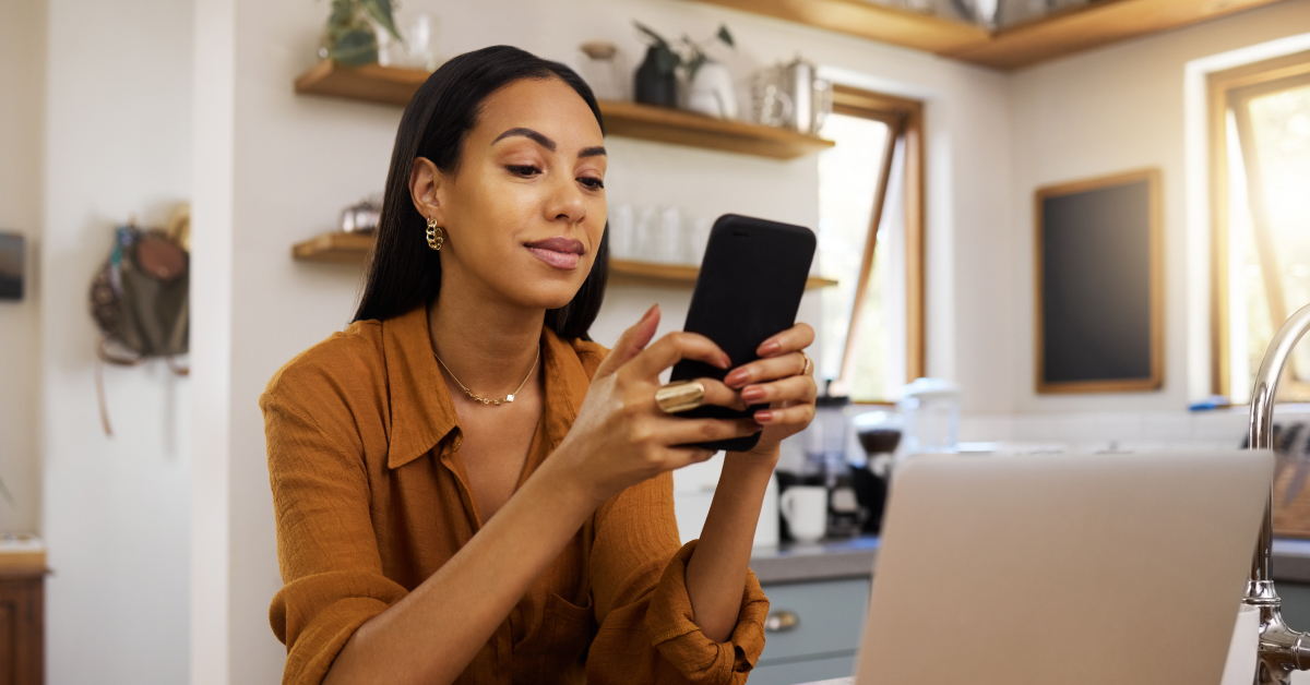 A woman is sitting in a cozy home office, smiling as she uses her smartphone. She appears relaxed and focused, with a laptop in front of her and a modern kitchen in the background. The setting suggests a comfortable and productive work-from-home environment, with natural light streaming through the windows. The image highlights the balance of professional and personal life in a home office setting.
