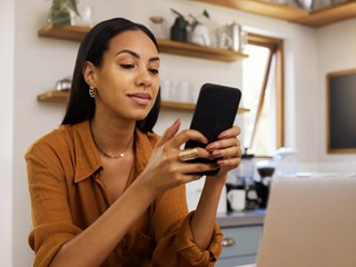 A woman is sitting in a cozy home office, smiling as she uses her smartphone. She appears relaxed and focused, with a laptop in front of her and a modern kitchen in the background. The setting suggests a comfortable and productive work-from-home environment, with natural light streaming through the windows. The image highlights the balance of professional and personal life in a home office setting.