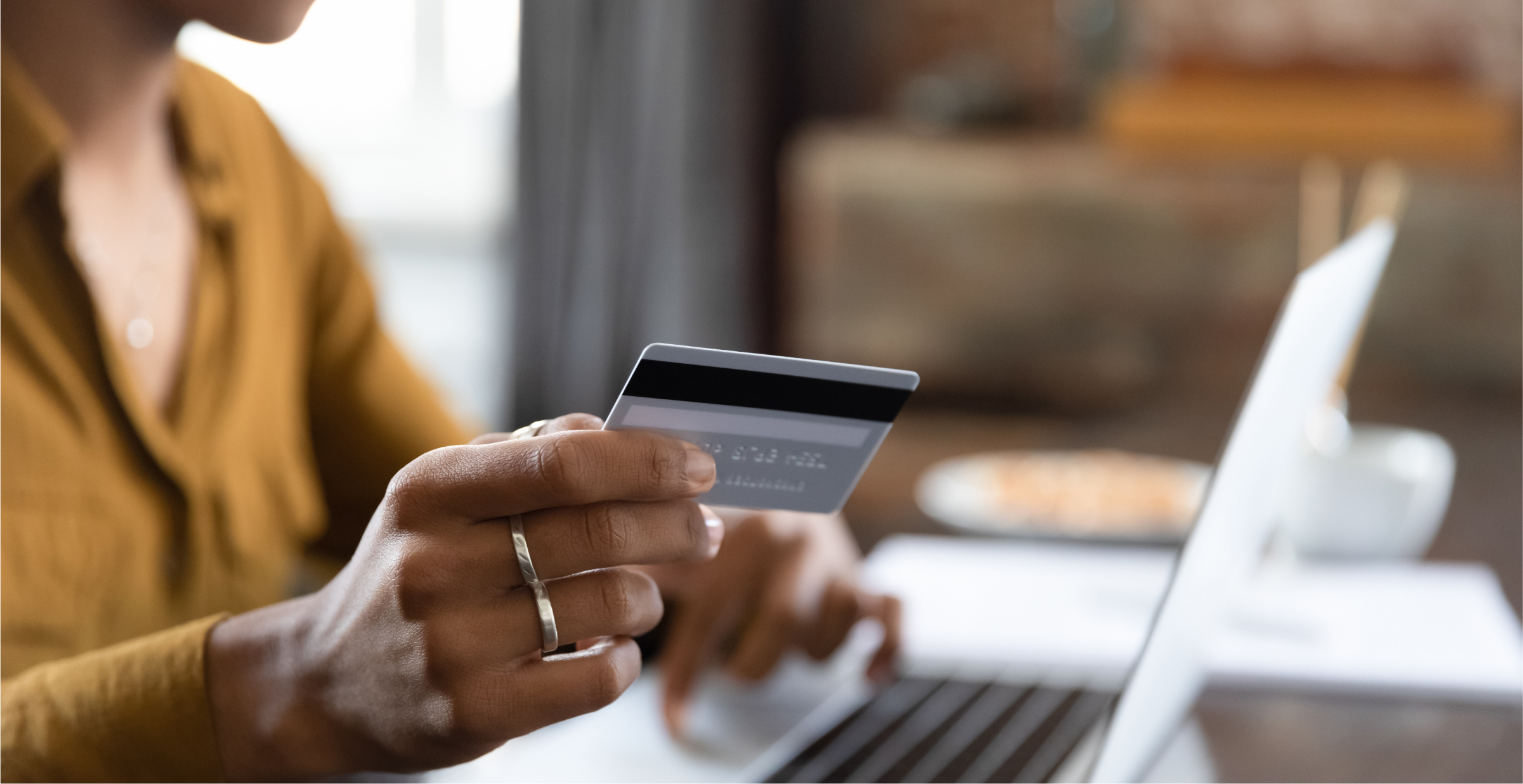 A close-up of a person holding a credit card while making an online purchase using a laptop. The person is typing with one hand and holding the card with the other, suggesting an online transaction or banking activity. The scene conveys the convenience and efficiency of digital financial services in a modern setting.