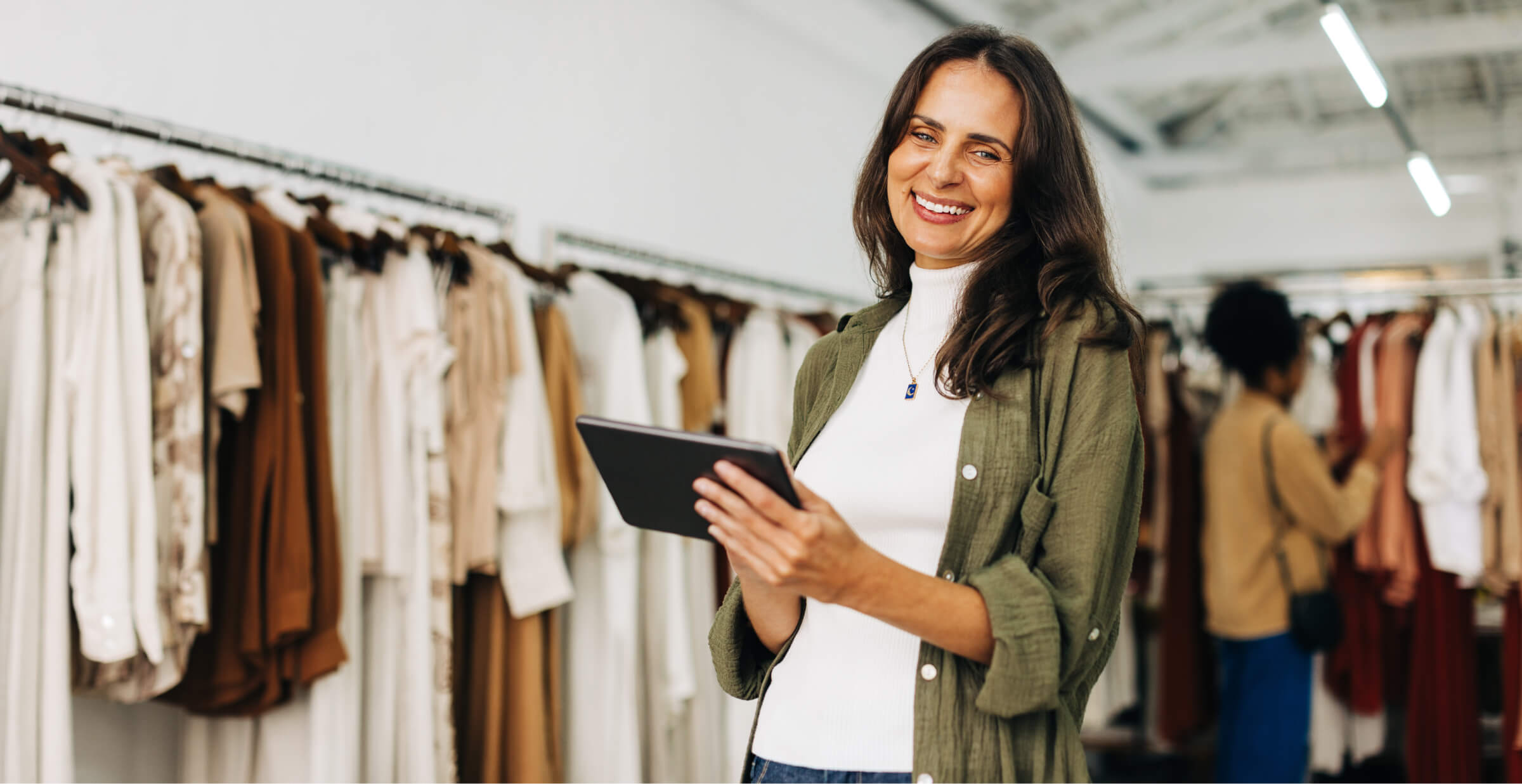 A smiling woman holding a tablet in a clothing store, standing in front of racks filled with neatly organized garments in neutral tones. She appears to be using the tablet for inventory or customer service. Another shopper is browsing the racks in the background.