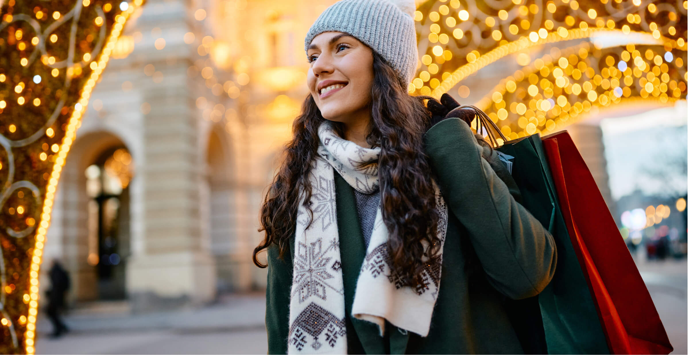 A smiling woman dressed in winter clothing, holding shopping bags while walking under festive holiday lights. She is wearing a knit hat, scarf, and coat, enjoying a seasonal shopping experience in a brightly lit, decorated urban setting.