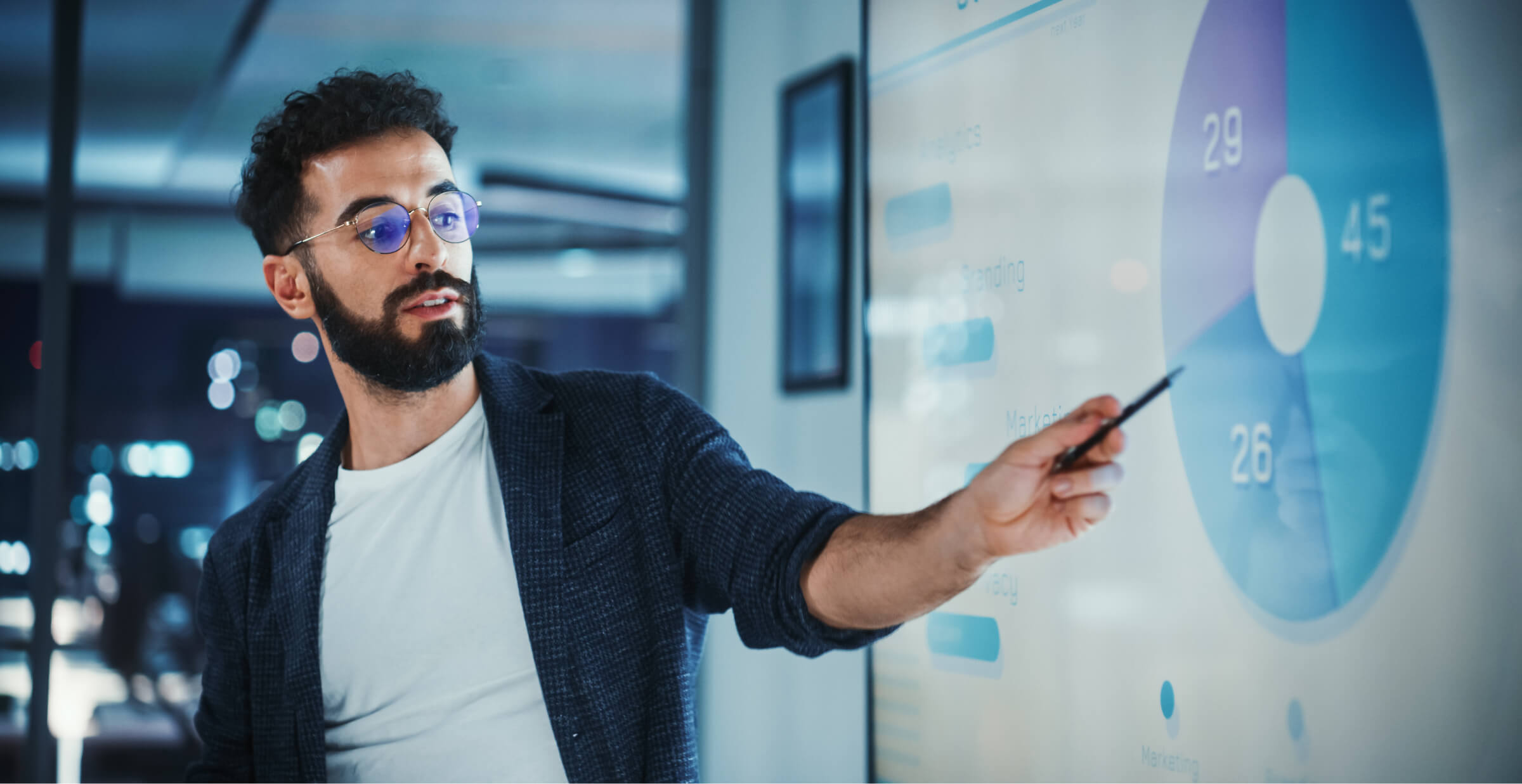A man with glasses presents data on a screen, pointing to a pie chart during a business meeting.