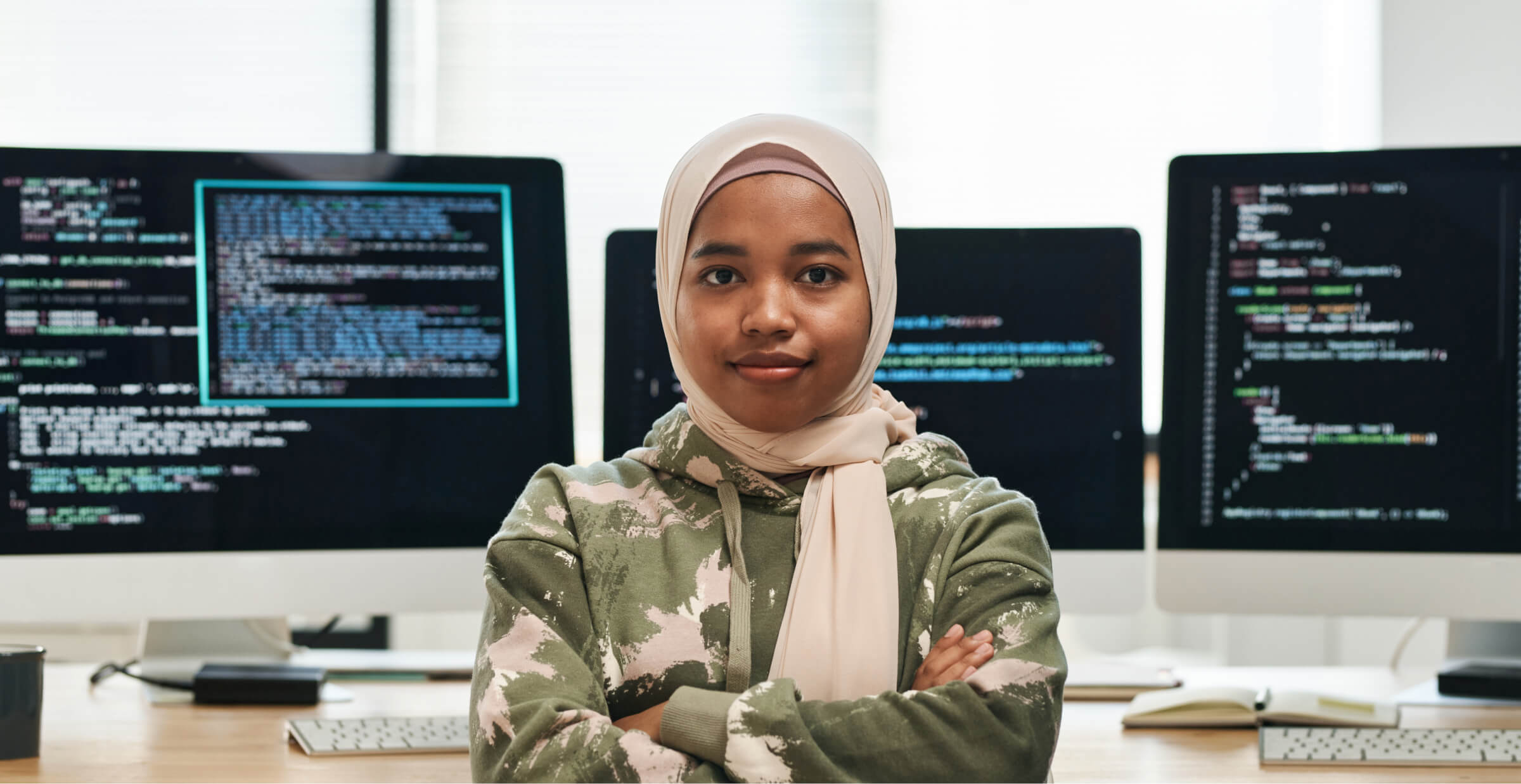 Confident software developer wearing a hijab, standing in front of computer screens with coding displayed