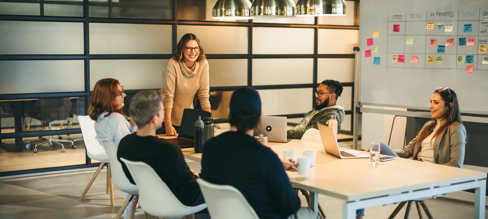 A group of colleagues sitting around a conference table, engaged in a collaborative meeting. A woman standing and smiling leads the discussion, while others listen and participate. The room has modern decor, with a whiteboard and laptops in use.