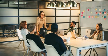 A group of colleagues sitting around a conference table, engaged in a collaborative meeting. A woman standing and smiling leads the discussion, while others listen and participate. The room has modern decor, with a whiteboard and laptops in use.