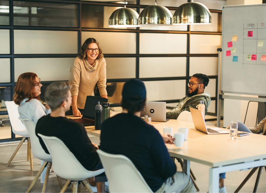 A group of colleagues sitting around a conference table, engaged in a collaborative meeting. A woman standing and smiling leads the discussion, while others listen and participate. The room has modern decor, with a whiteboard and laptops in use.