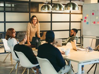A group of colleagues sitting around a conference table, engaged in a collaborative meeting. A woman standing and smiling leads the discussion, while others listen and participate. The room has modern decor, with a whiteboard and laptops in use.