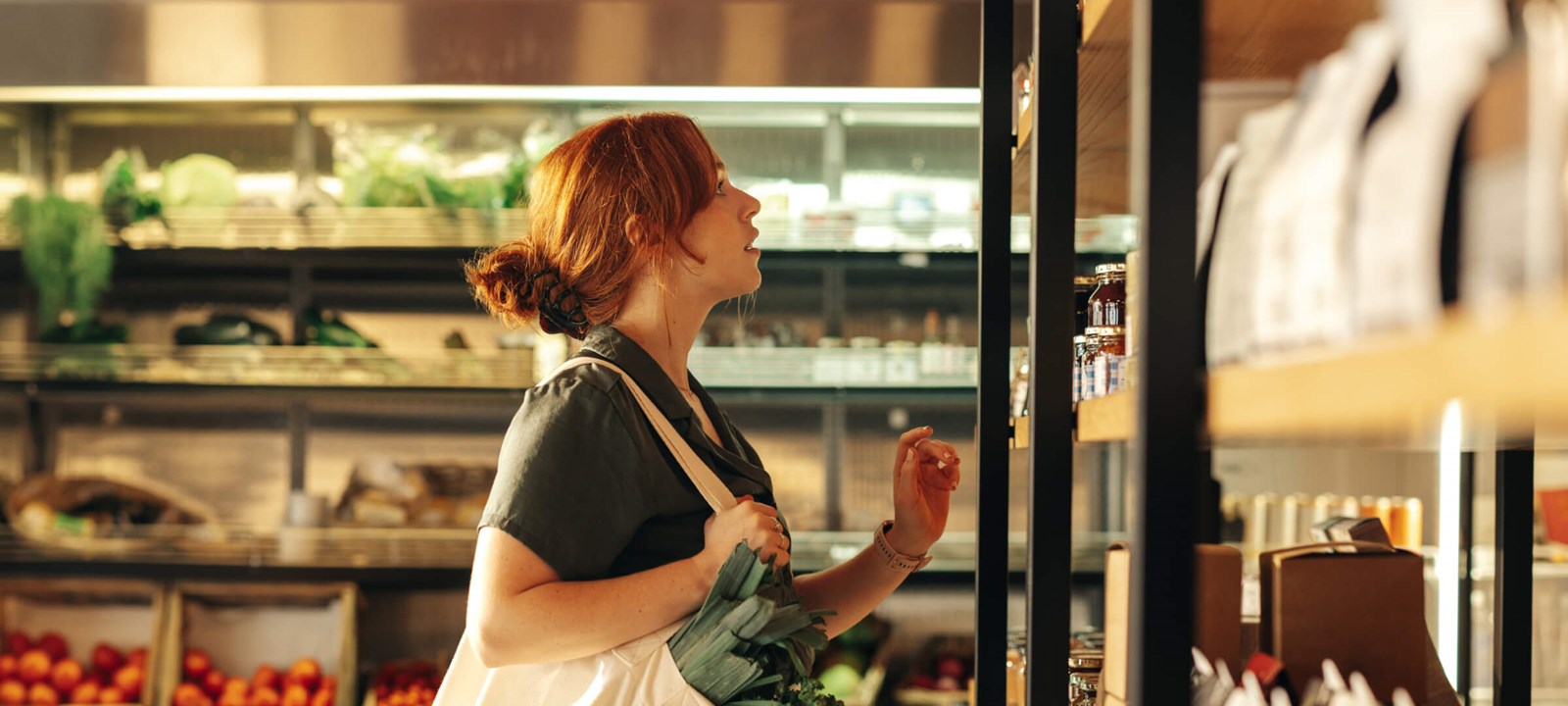 Red-haired woman browsing organic products in a deli store, carrying a tote bag filled with leafy greens.