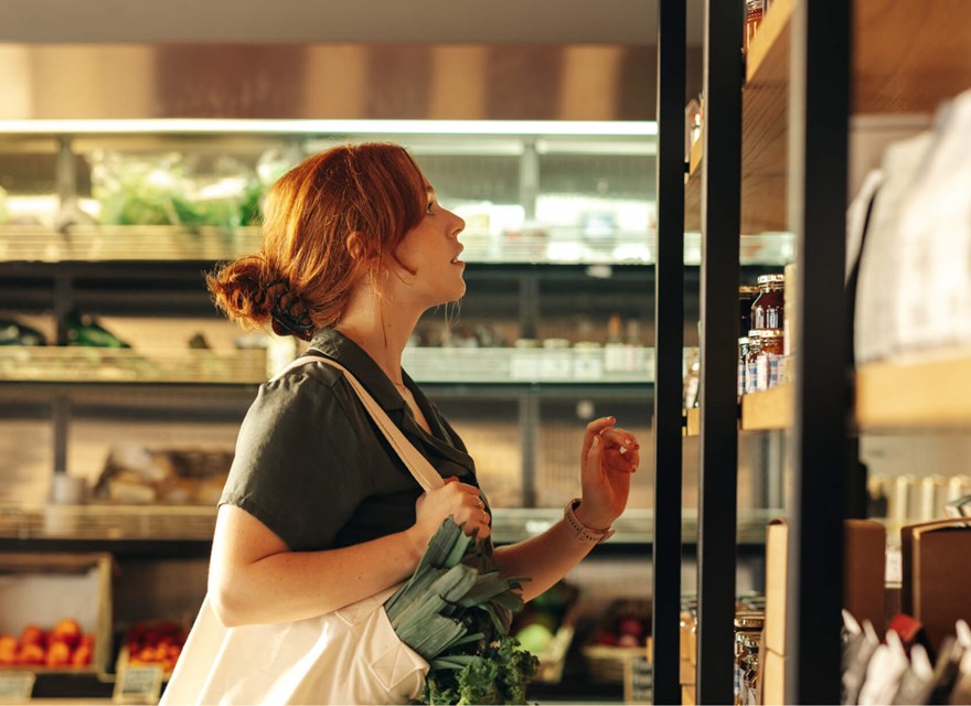 Red-haired woman browsing organic products in a deli store, carrying a tote bag filled with leafy greens.