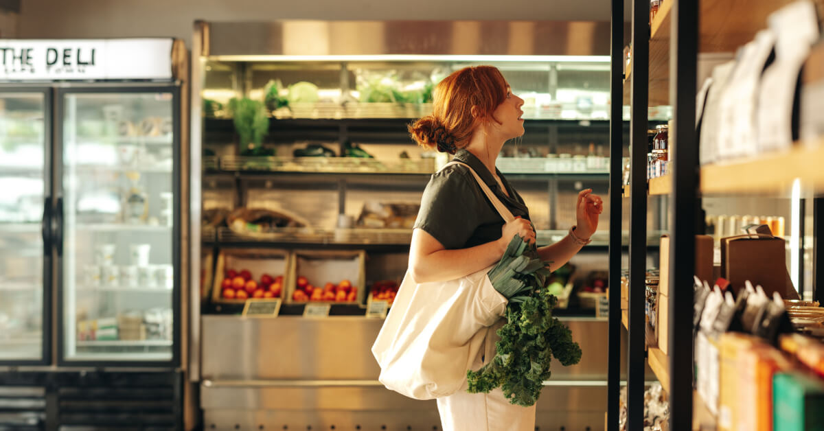 Red-haired woman browsing organic products in a deli store, carrying a tote bag filled with leafy greens.