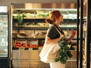 Red-haired woman browsing organic products in a deli store, carrying a tote bag filled with leafy greens.