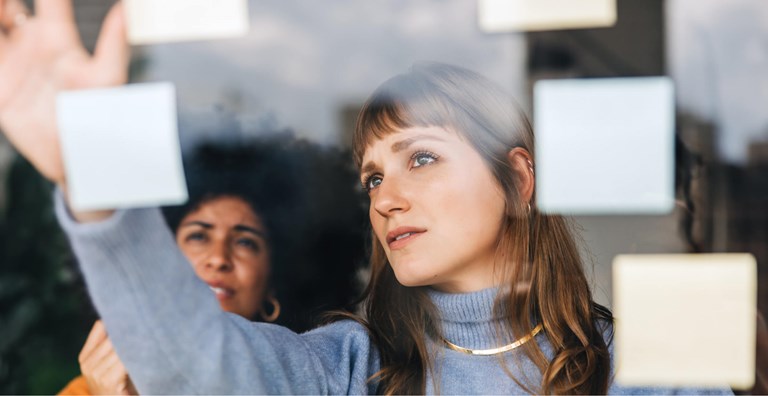 Two women collaborating in a brainstorming session, placing sticky notes on a glass wall. The woman in the foreground is focused as she arranges the notes, while the second woman observes from behind. The scene conveys teamwork and creative problem-solving.