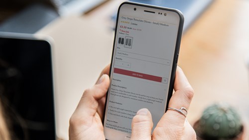 A person holding a smartphone, viewing a product page for Grey Stripe Thinsulate Gloves. The screen shows the product description, price, and purchasing options. In the background, a laptop and a small potted cactus are visible, suggesting a casual or work-from-home environment.