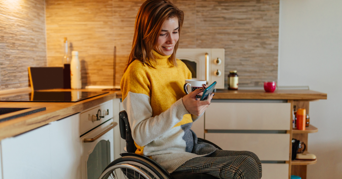 A woman sitting in a wheelchair in a modern kitchen, wearing a yellow and gray sweater, holds a coffee mug in one hand and looks at her mobile phone with a smile. The background shows kitchen countertops, cabinets, and appliances, giving a cozy and relaxed atmosphere.