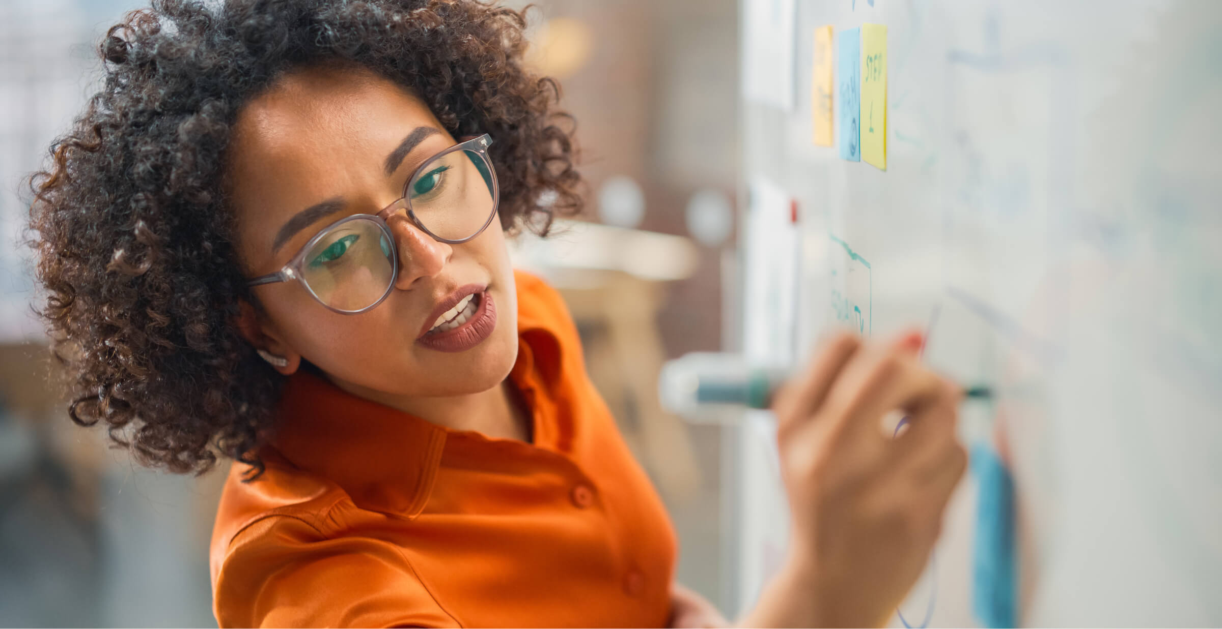 A focused woman with curly hair and glasses is writing on a whiteboard during a brainstorming or planning session. Colorful sticky notes and diagrams are visible on the board, suggesting an organized and collaborative work process. The image captures the energy and creativity of a modern professional environment, highlighting problem-solving and strategic thinking.