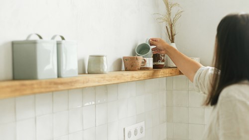 A person is seen from behind while placing a light blue ceramic cup on a wooden shelf in a minimalistic kitchen. The shelf is decorated with various ceramic mugs, jars, and a vase with dried plants. The walls have a light tone, and the tiled backsplash is white. The overall decor exudes a clean, modern, and cozy aesthetic.