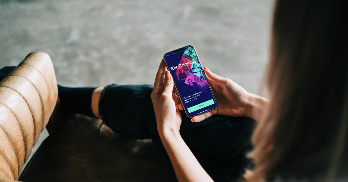 A person sitting in a chair, holding a smartphone and viewing an app screen for The Telegraph. The screen displays vibrant, colorful graphics and a 'Get Started' button, suggesting the user is on a welcome or sign-up page.