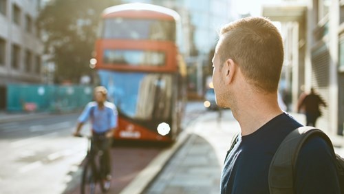 A man with a backpack stands on a city street, looking toward an approaching red double-decker bus. A cyclist rides past on the street, with blurred city buildings in the background, suggesting an urban commuting scene.