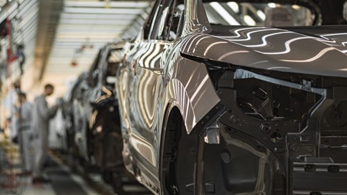 An automotive car assembly line in a factory with several vehicles in various stages of construction and workers blurred in the background.
