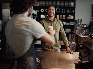 A customer at a café counter smiles as they receive a takeaway coffee cup from a barista. The customer, dressed in a light green button-up shirt, stands on the other side of the counter. The café background features shelves with various items, such as mugs and coffee bags. The atmosphere is warm and inviting.