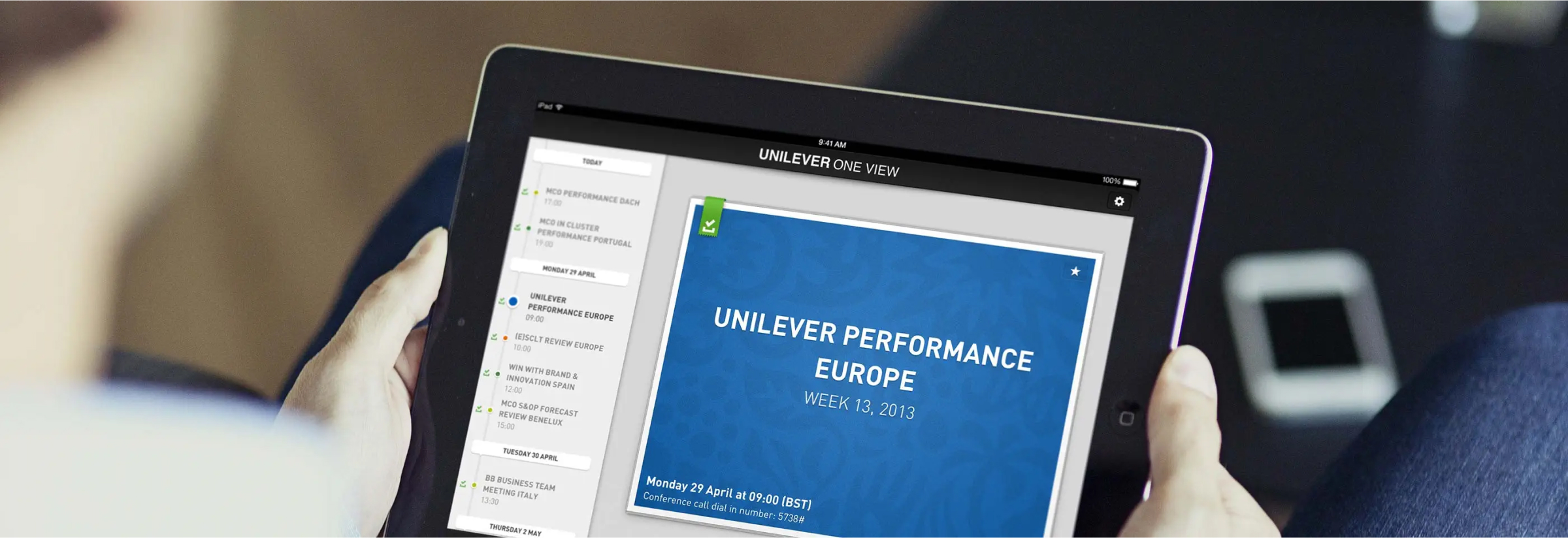 Two hands hold a tablet displaying a presentation slide titled "Unilever Performance Europe Week 12, 2013." In the top left of the presentation slide, there’s a green checkmark icon, possibly indicating the content is downloaded from the cloud. The screen also shows a side panel with a list of items, sorted by date, where each item has a green checkmark as well.