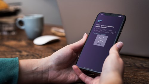 A person holding an iPhone displaying a log in screen. The FaceID functionality of iOS is displayed, together with a heading that reads “Log in with your Bankline Mobile Keycode”, followed by six empty number fields. Below there’s a text link saying “Forgotten your Keycode?”.