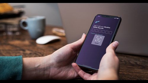 A person holding an iPhone displaying a log in screen. The FaceID functionality of iOS is displayed, together with a heading that reads “Log in with your Bankline Mobile Keycode”, followed by six empty number fields. Below there’s a text link saying “Forgotten your Keycode?”.