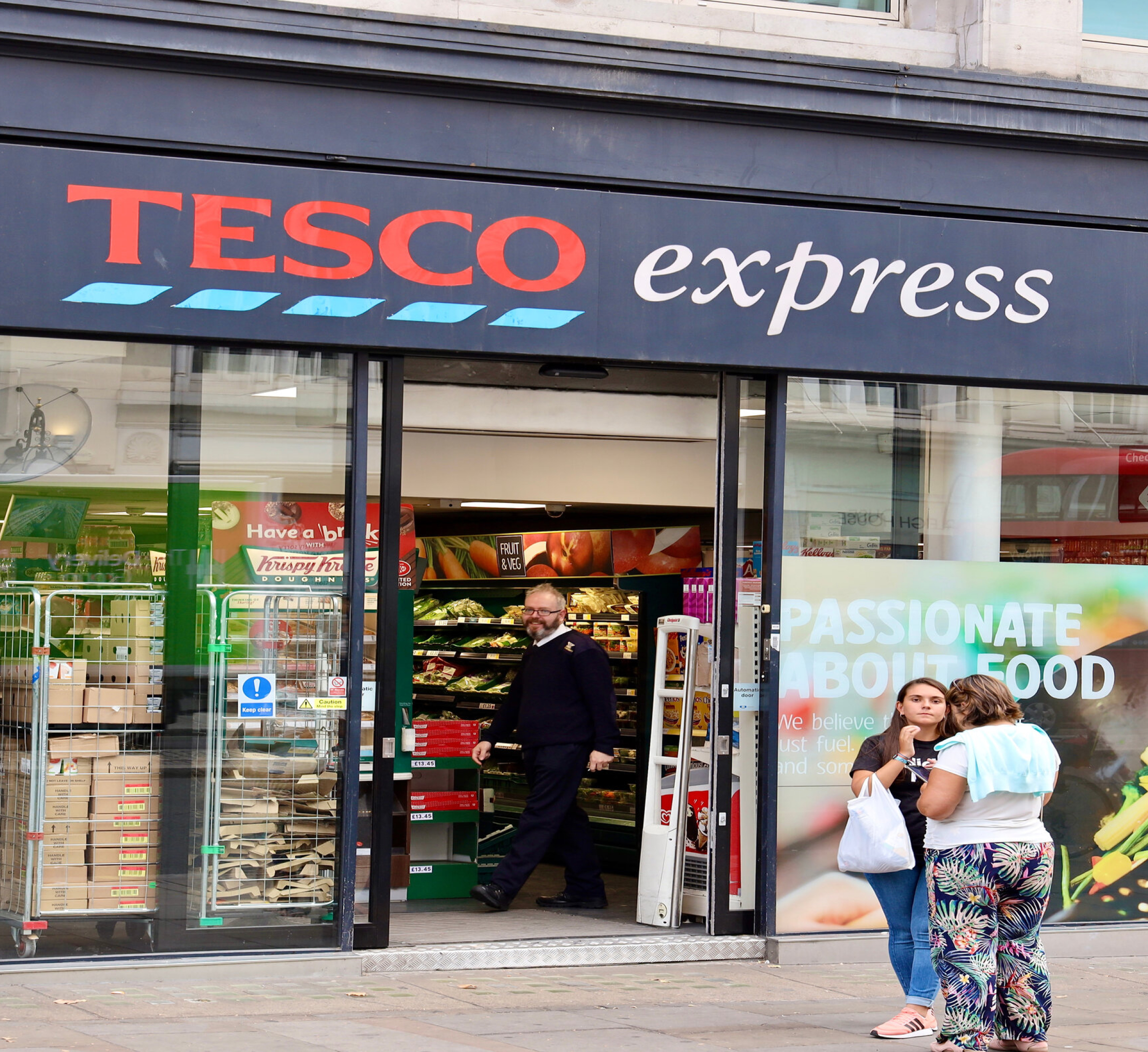 A Tesco Express storefront with a glass entrance, showing a man walking out of the store and two women standing outside, one holding a shopping bag. The store's interior displays fresh produce and other grocery items, and a large sign promoting the brand's passion for food is visible on the right.