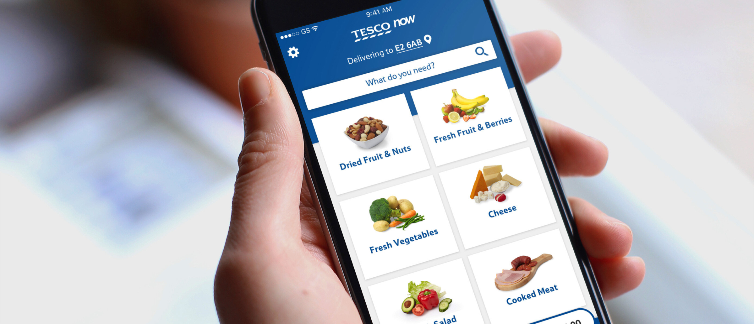 A person holding a smartphone displaying the Tesco Now app. The app screen shows various grocery categories such as dried fruit & nuts, fresh vegetables, cheese, and cooked meat, with an easy-to-use interface for selecting items to order for delivery.