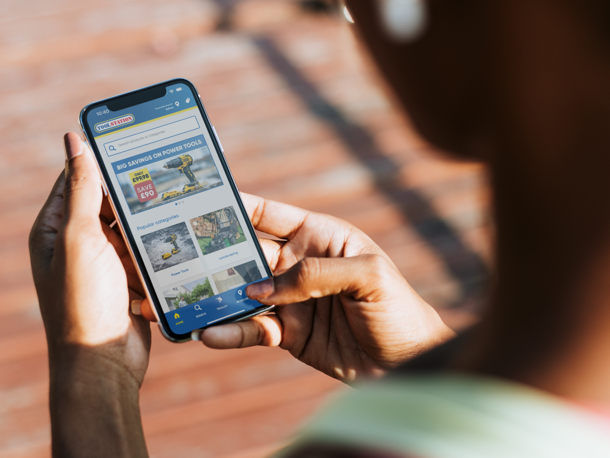 A person holding a smartphone and browsing the Toolstation website, viewing power tools and popular categories like landscaping and gardening. The screen displays a promotion for power tool savings, with a clear, user-friendly interface.