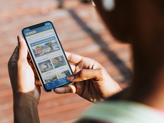 A person holding a smartphone and browsing the Toolstation website, viewing power tools and popular categories like landscaping and gardening. The screen displays a promotion for power tool savings, with a clear, user-friendly interface.